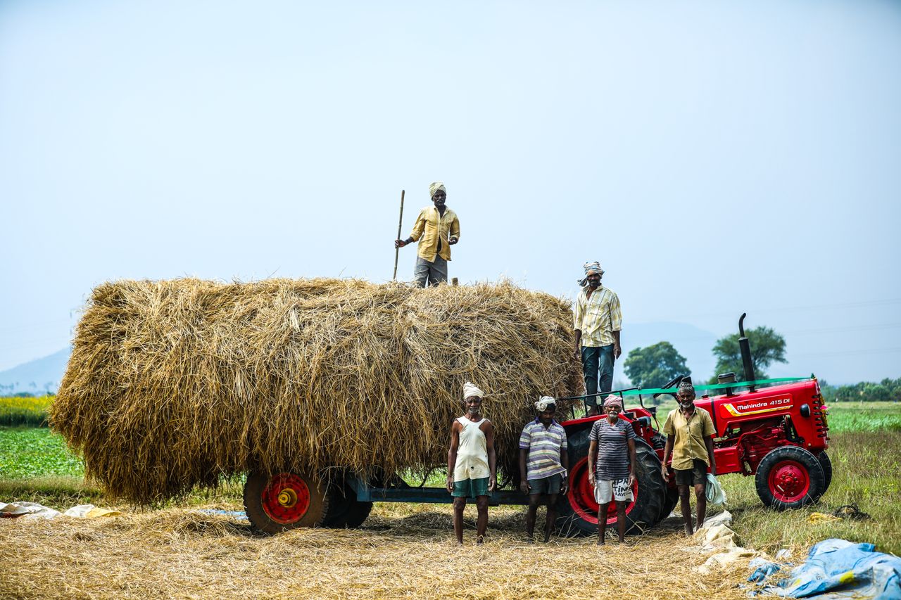 Indian farmers posing with a haystack on a tractor in a countryside field, showcasing rural agriculture.